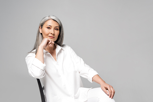 serious asian woman with grey hair sitting on chair isolated on grey