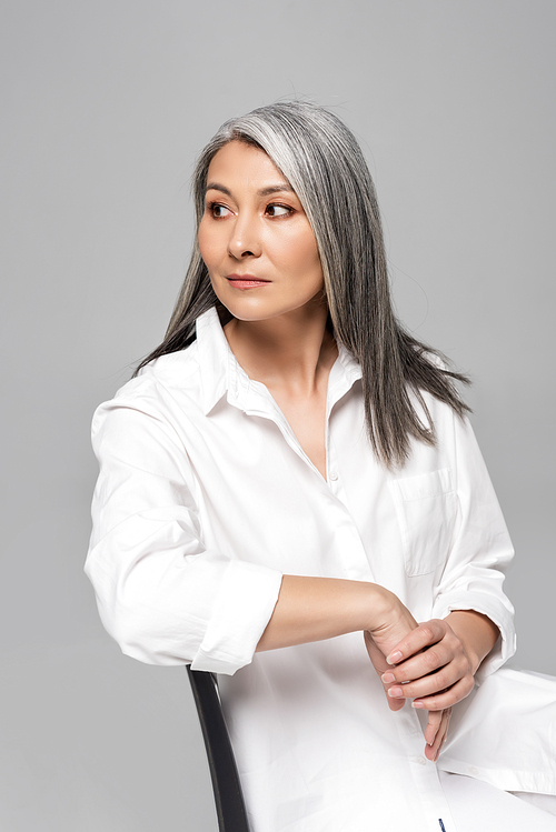 confident asian woman with grey hair sitting on chair isolated on grey
