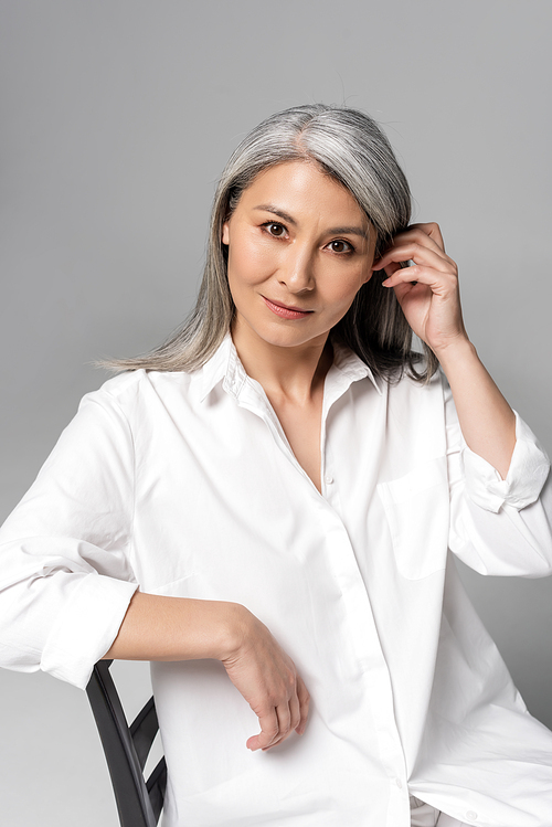 thoughtful asian woman with grey hair sitting on chair isolated on grey