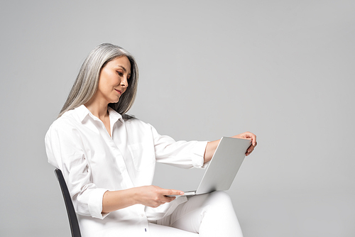 adult asian woman with grey hair sitting on chair and using laptop isolated on grey