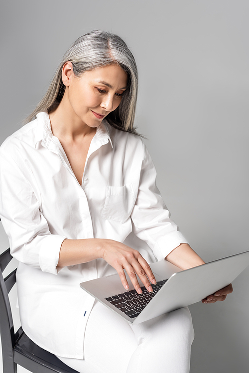 smiling asian woman with grey hair sitting on chair and using laptop on grey
