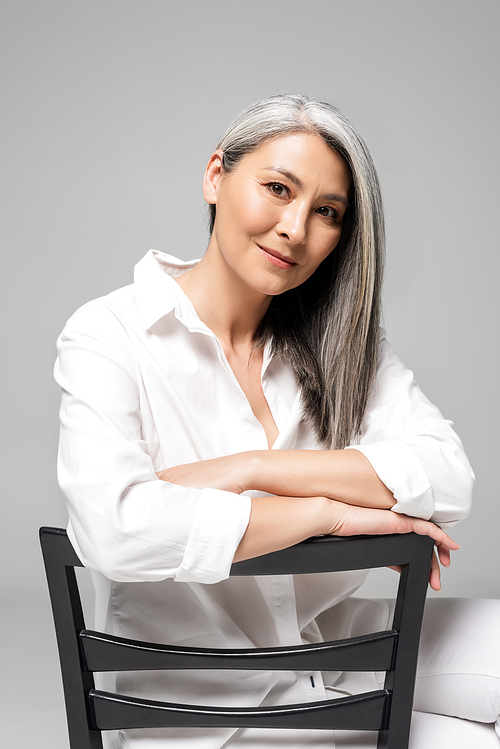 smiling asian woman with grey hair sitting on chair isolated on grey