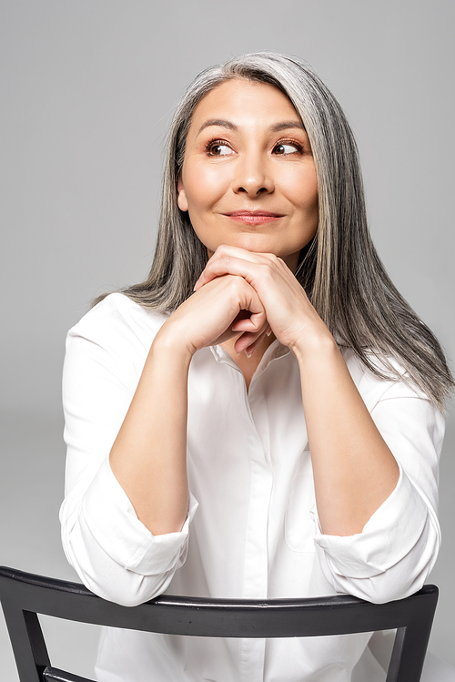 positive pensive asian woman with grey hair sitting on chair isolated on grey