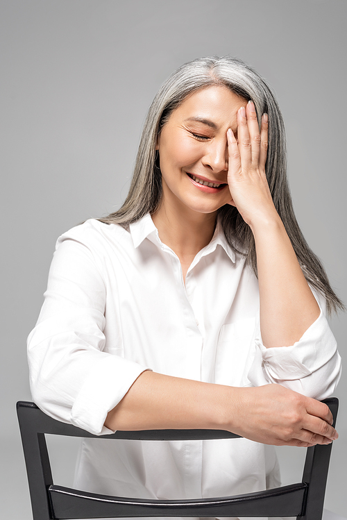 beautiful laughing asian woman with grey hair sitting on chair isolated on grey