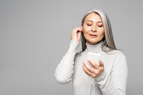 asian woman in turtleneck with grey hair listening music with earphones and smartphone isolated on grey