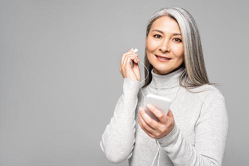 happy asian woman in turtleneck with grey hair listening music with earphones and smartphone isolated on grey