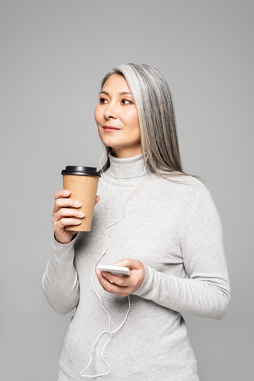 beautiful asian woman in turtleneck with coffee to go listening music with earphones and smartphone isolated on grey