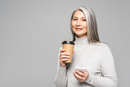 happy asian woman in turtleneck with coffee to go listening music with earphones and smartphone isolated on grey