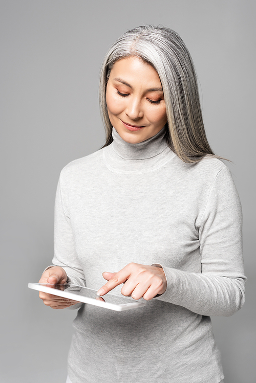 happy adult asian woman in turtleneck using digital tablet isolated on grey
