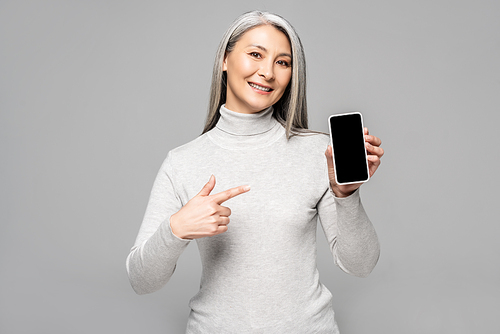 happy asian woman in turtleneck with grey hair pointing at smartphone with blank screen isolated on grey