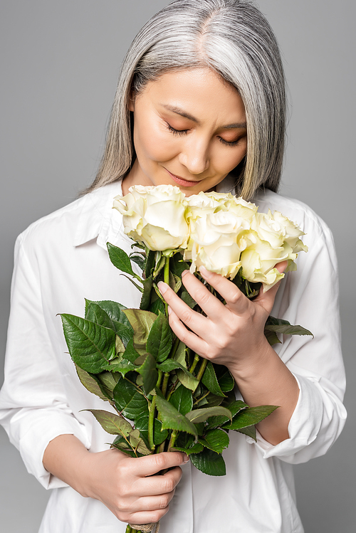 adult tender asian woman with grey hair and closed eyes holding bouquet of white roses isolated on grey