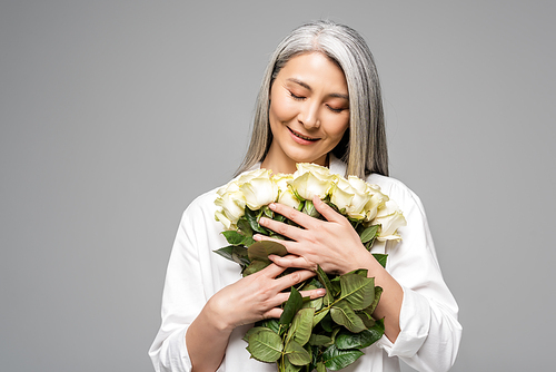 thankful asian woman with grey hair holding bouquet of white roses isolated on grey