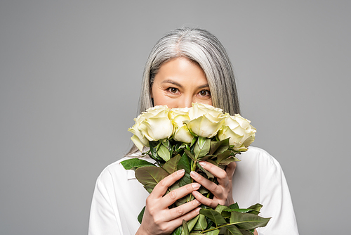 positive asian woman with grey hair holding bouquet of white roses in front of face isolated on grey