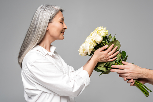 positive adult asian woman with grey hair in white shirt taking bouquet of white roses from man isolated on grey