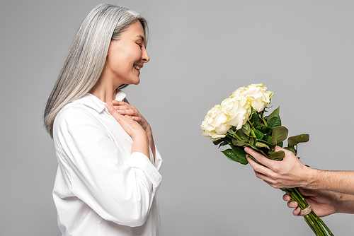 happy adult asian woman with grey hair in white shirt looking at bouquet of white roses from man isolated on grey