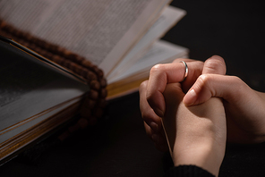 cropped view of woman praying near holy bible with rosary in dark