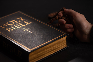 cropped view of woman holding rosary near holy bible in dark with sunlight