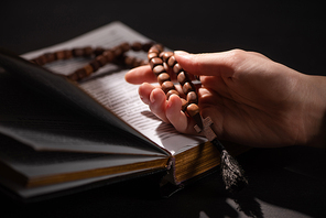 cropped view of woman holding rosary with cross near open holy bible in dark with sunlight