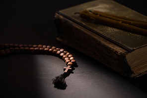 selective focus of holy bible with candles and rosary with cross in dark with sunlight