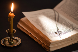 church candle in candlestick burning near bible with catholic cross in dark with sunlight