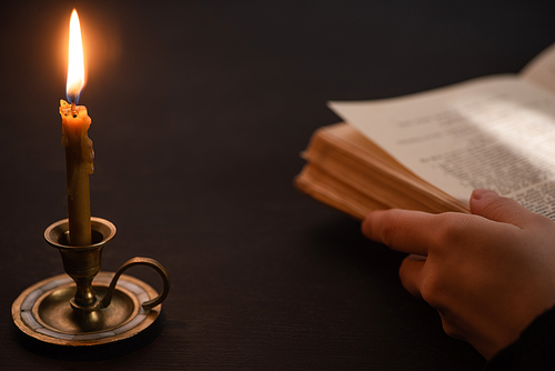 cropped view of woman holding holy bible near burning candle in dark