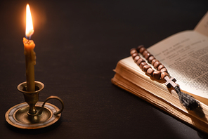 church candle in candlestick burning near bible with catholic rosary in dark