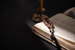 selective focus of candlestick near bible with catholic rosary in dark with sunlight