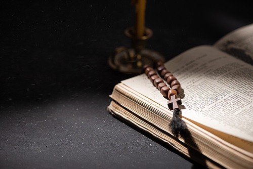 selective focus of candlestick near bible with catholic rosary in dark with sunlight
