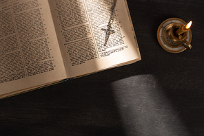 top view of open holy bible with cross near candle on dark background with sunlight