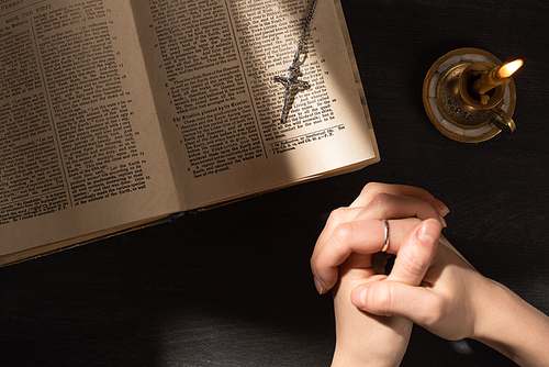 cropped view of woman praying near open holy bible with cross on dark background with sunlight