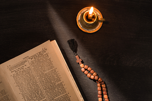 top view of open holy bible with rosary and candle on dark background with sunlight