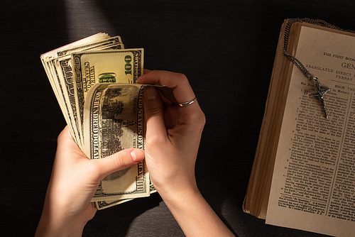 cropped view of woman counting money near holy bible with cross on dark background with sunlight