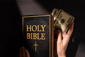 cropped view of woman taking money out of holy bible on dark background with sunlight