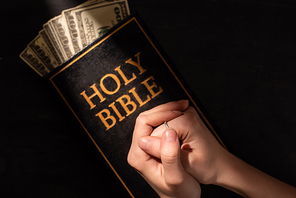 cropped view of woman praying on holy bible with money on dark background with sunlight