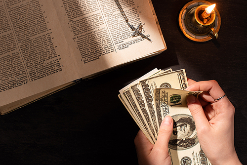 cropped view of woman counting money near holy bible with cross and candle on dark background with sunlight