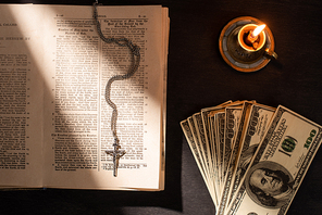 top view of holy bible with money, cross and candle on dark background with sunlight