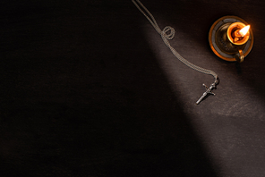 top view of burning candle and cross on dark background with sunlight