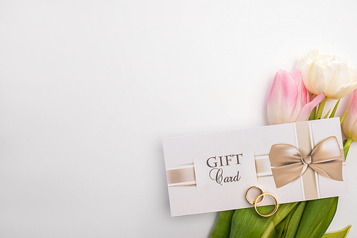 Top view of gift card, wedding rings and tulips on white background