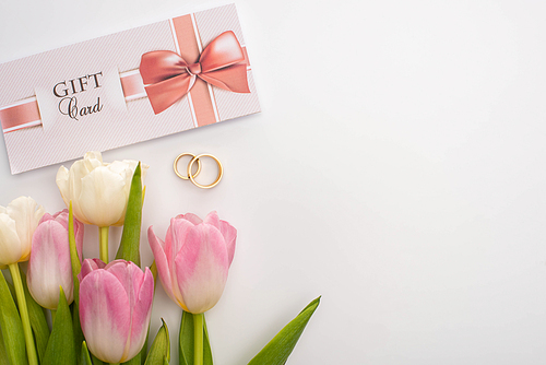 Top view of gift card near flowers and wedding rings on white background