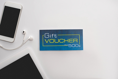 Top view of gift voucher near smartphone, digital tablet and earphones on white surface