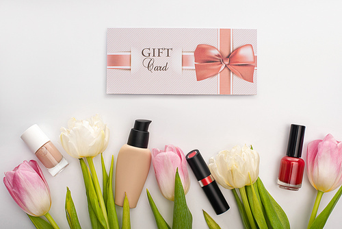 Top view of gift card near decorative cosmetics and flowers on white surface