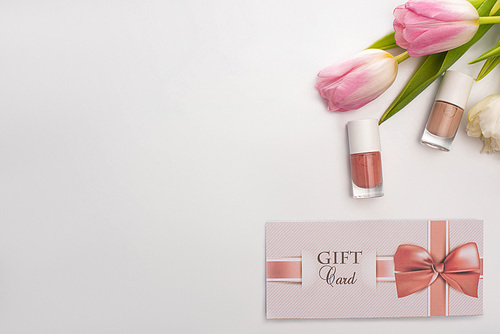 Top view of gift card near nail polishes and flowers on white surface