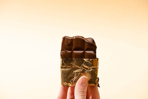cropped view of woman holding delicious milk chocolate bar in golden foil isolated on beige
