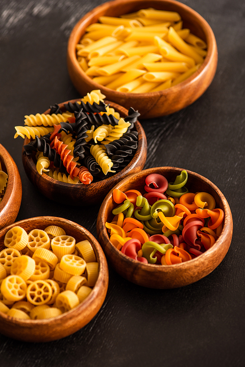 selective focus of various raw Italian pasta in wooden bowls on black background