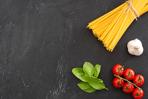 top view of raw Italian spaghetti with basil leaves, tomatoes and garlic on black background