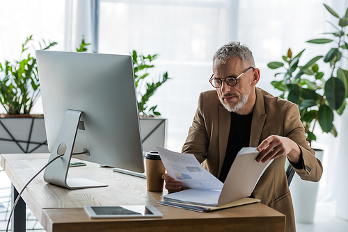 selective focus of bearded businessman in glasses looking at document near coffee to go and computer monitor in office