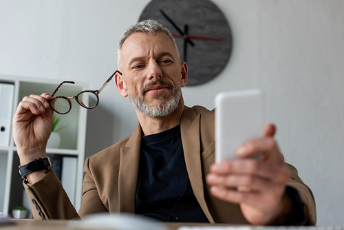 selective focus of businessman holding glasses and looking at smartphone in office