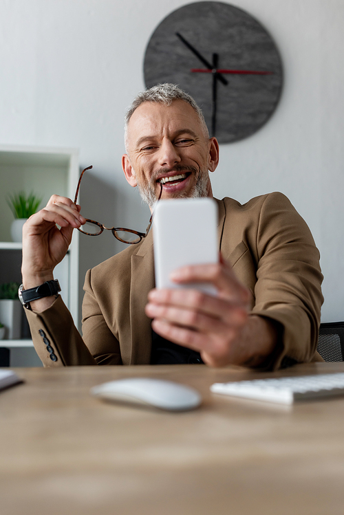 selective focus of happy businessman holding glasses and looking at smartphone in office