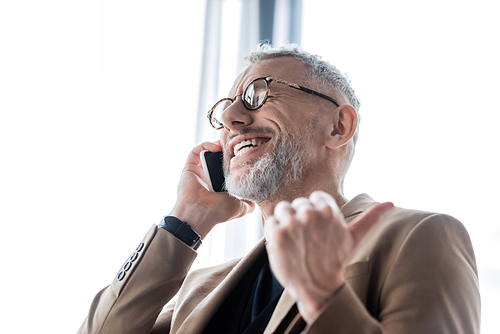 selective focus of happy and bearded businessman talking on smartphone and showing thumb up