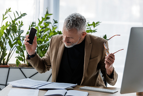 bearded businessman holding glasses and smartphone with blank screen while looking at documents near computer monitor